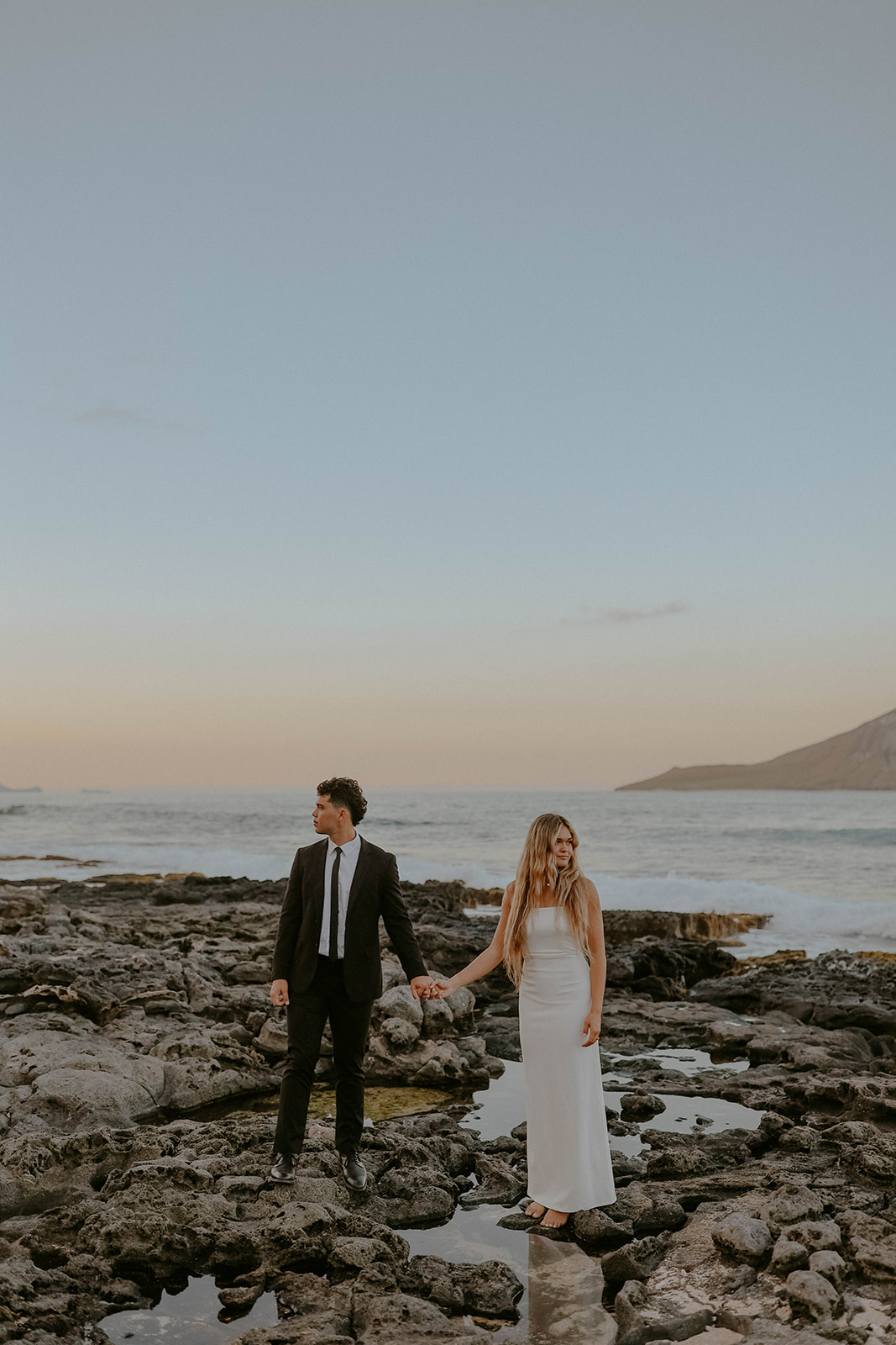 a bride and groom stand on rocks next to the ocean at their oahu elopement