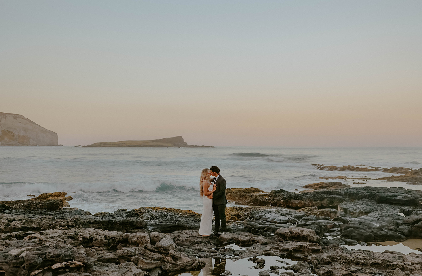 a bride and groom stand on rocks next to the ocean at their oahu elopement

