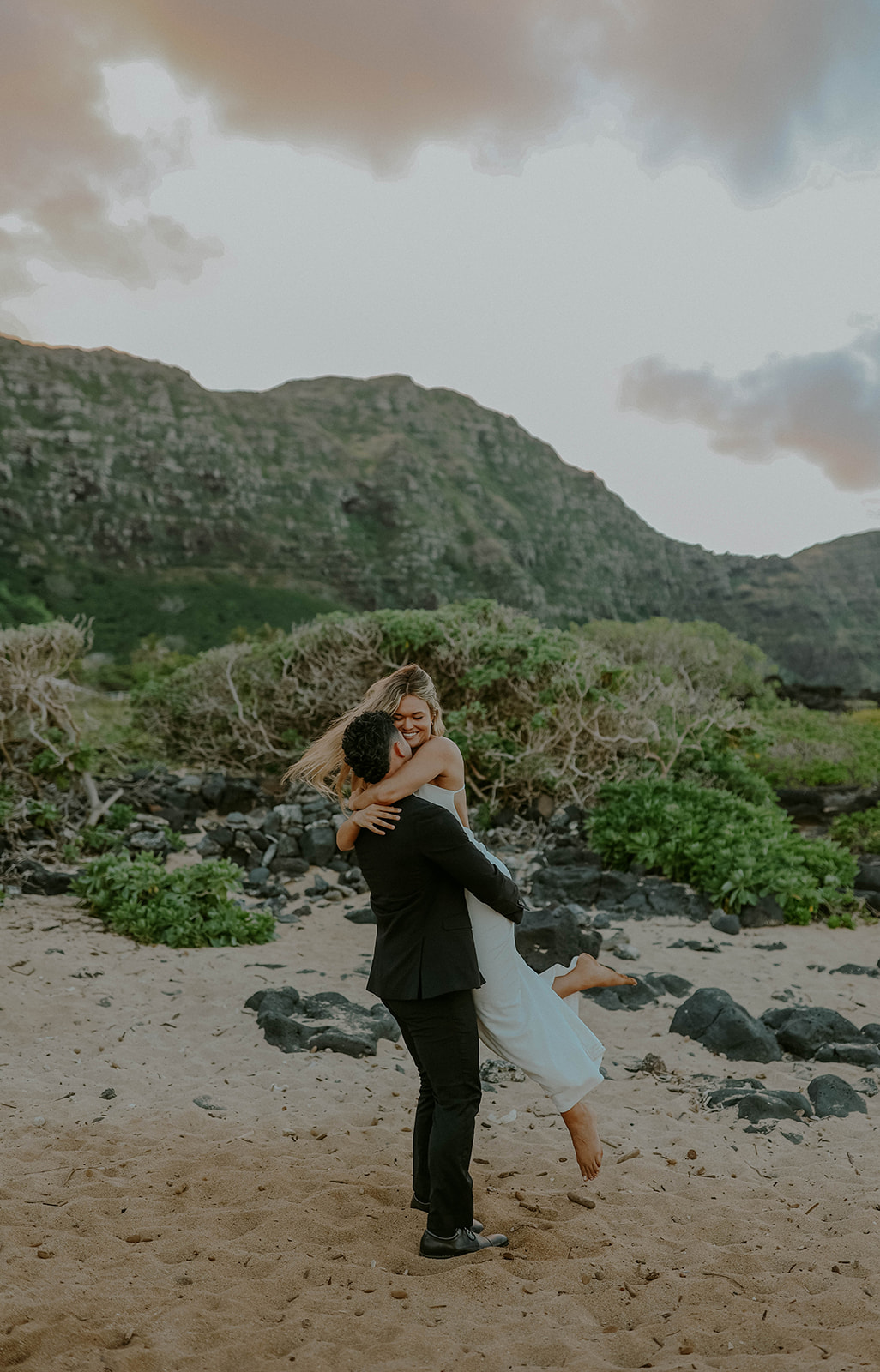 a groom picks up his bride during their oahu elopement while they pose in the sand on a beach with lush cliffs in the background