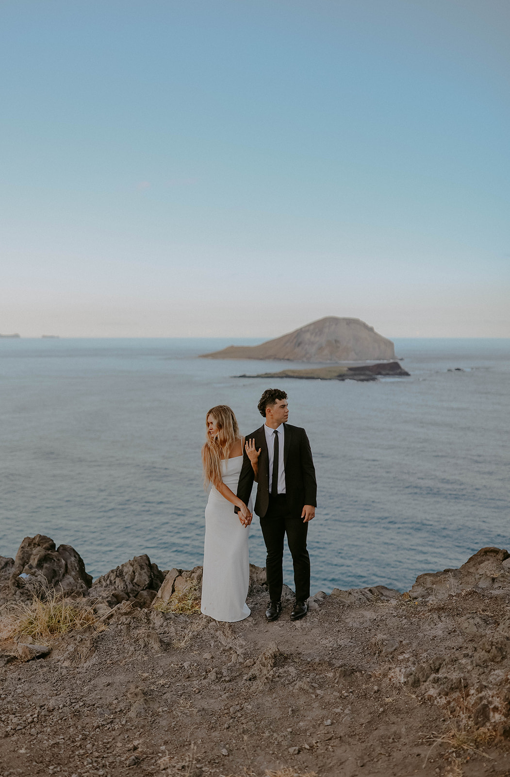 a bride and groom dressed in wedding day outfits pose on a cliff overlooking the ocean during their elopement in oahu hawaii