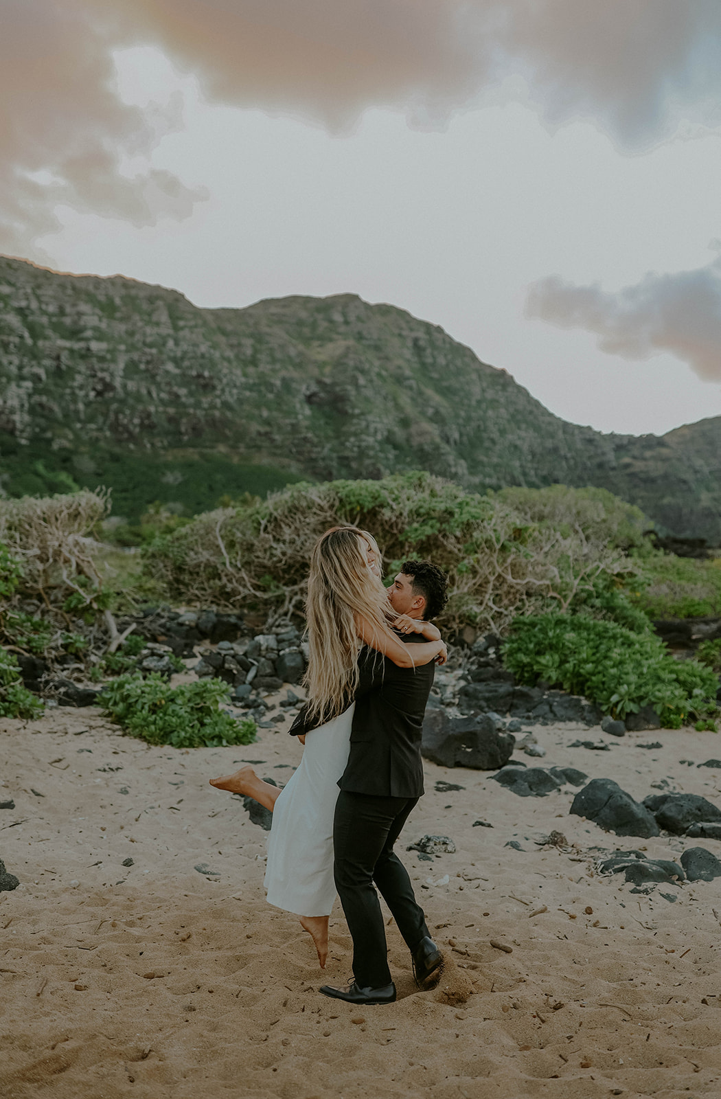 a groom picks up his bride during their oahu elopement while they pose in the sand on a beach with lush cliffs in the background